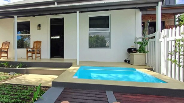 Ein modernes Haus mit einer überdachten Terrasse, zwei Holzstühlen, einem Schwimmbecken und einem eingezäunten Garten.