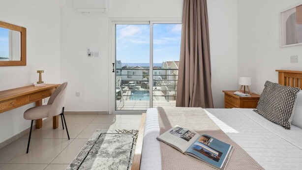 Un dormitorio moderno con cama, escritorio, silla, mesita de noche y vistas a edificios a través de un ventanal.