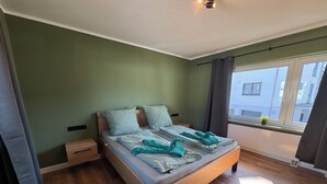 Une chambre avec un lit en bois, des murs verts, une fenêtre donnant sur des immeubles et une petite table de chevet.