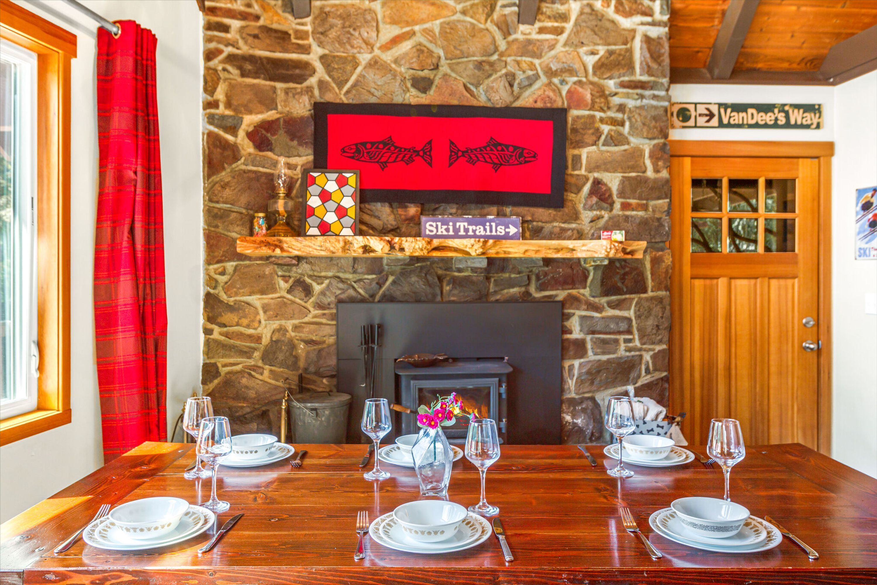 Una mesa de comedor dispuesta con platos blancos, vasos, cubiertos, un florero con flores y una chimenea con un fondo de pared de piedra.