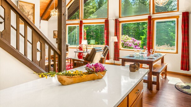A spacious room with a wooden staircase, large windows with red curtains, a wooden dining table, and a flower vase on the counter. - Cottonwood Chalet in Packwood (Packwood)