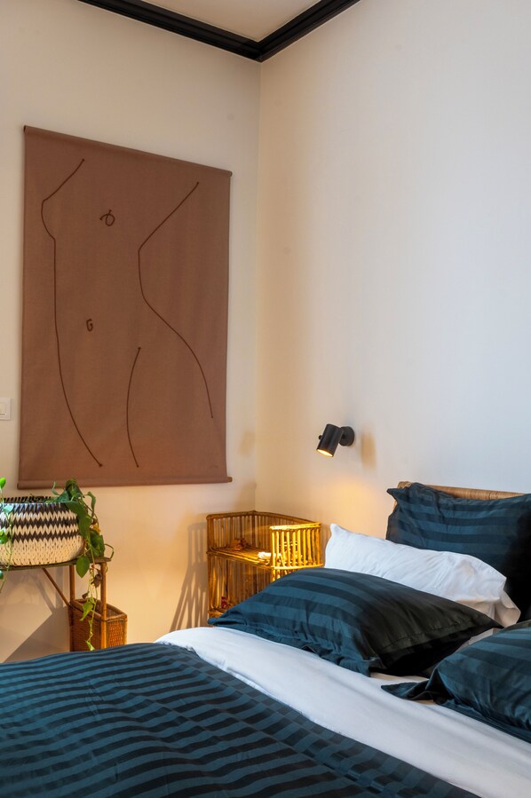 A bedroom with a large abstract painting, a bedside lamp, a wicker side table, and a bed with dark blue bedding.