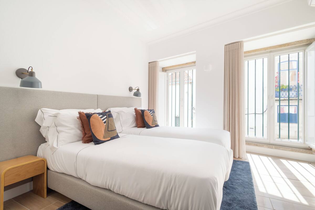 Cama doble con ropa de cama blanca y dos almohadas, una mesita de noche, una lámpara de pared y una ventana con cortinas.