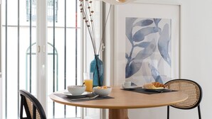 A dining area with a round wooden table, two wicker chairs, a vase with dried branches, a glass of orange juice, a bowl of fruit, a plate with croissants, and a framed abstract painting on the wall. - Lapa Diplomatic Suite&Flat by FeelLisboa (Lisbon)