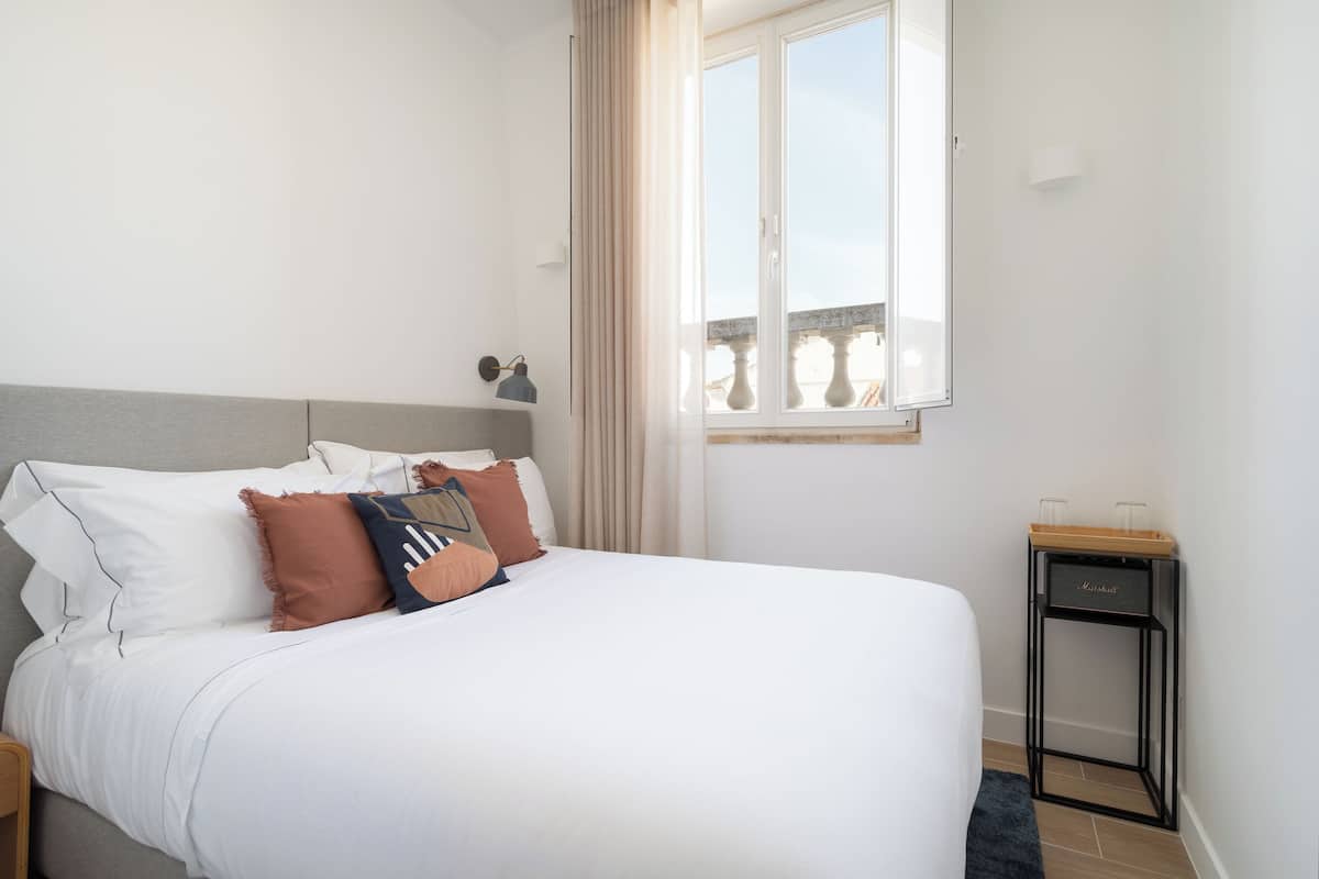 Una cama bien tendida con ropa de cama blanca y almohadas decorativas, una mesita de noche con lámpara y una ventana con cortinas.