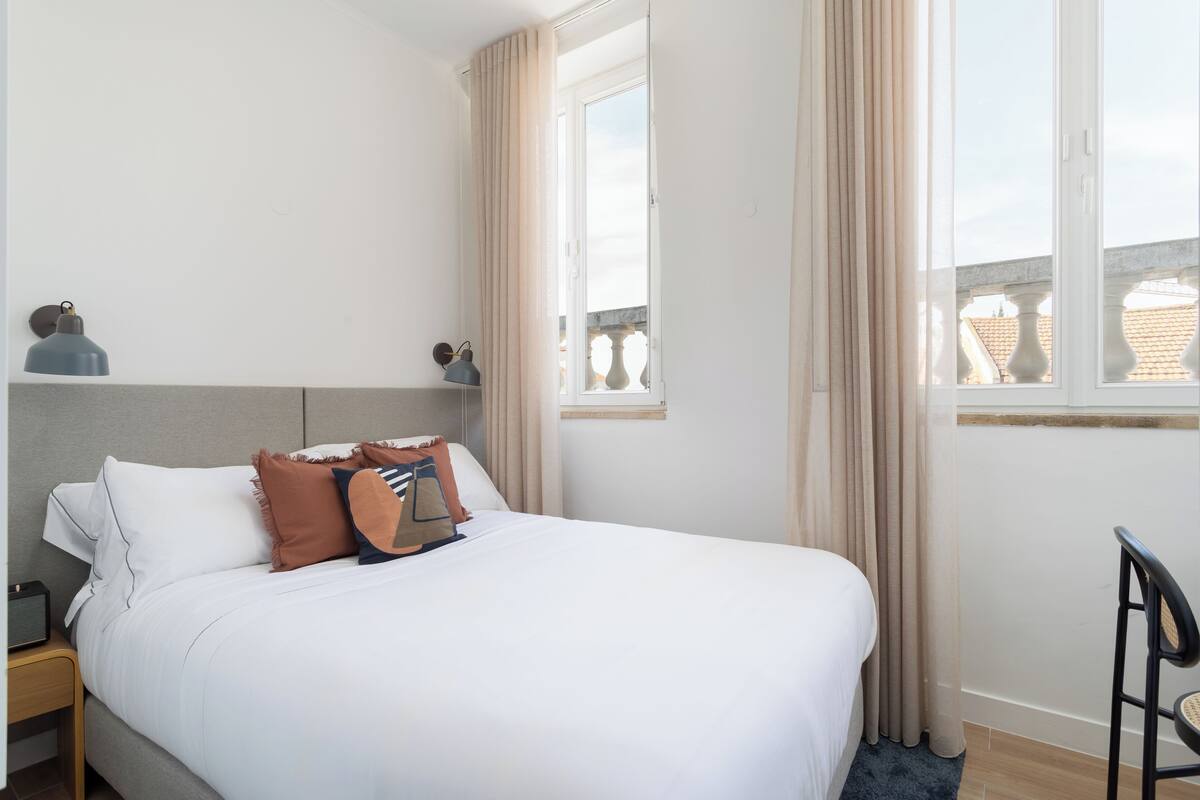 Una cama bien tendida con sábanas blancas y algunos almohadones decorativos, una mesita de noche con una lámpara y una ventana con cortinas.