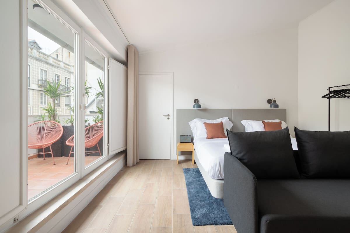Una habitación de hotel moderna con un ventanal grande, una cama con ropa oscura, un tapete azul y una mesita de noche con una lámpara.