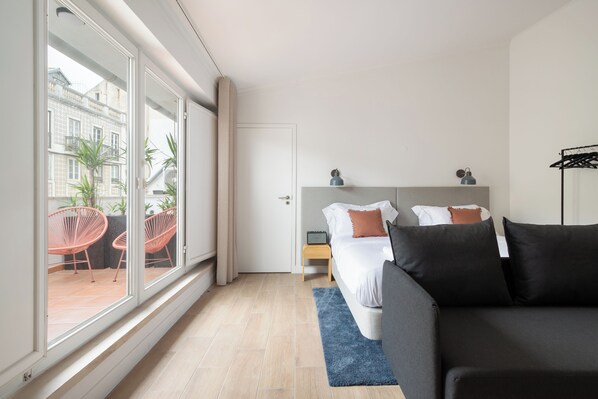 A modern hotel room with a large window, a bed with dark bedding, a blue rug, and a small bedside table with a lamp. - Lapa Diplomatic Suite&Flat by FeelLisboa (Lisbon)