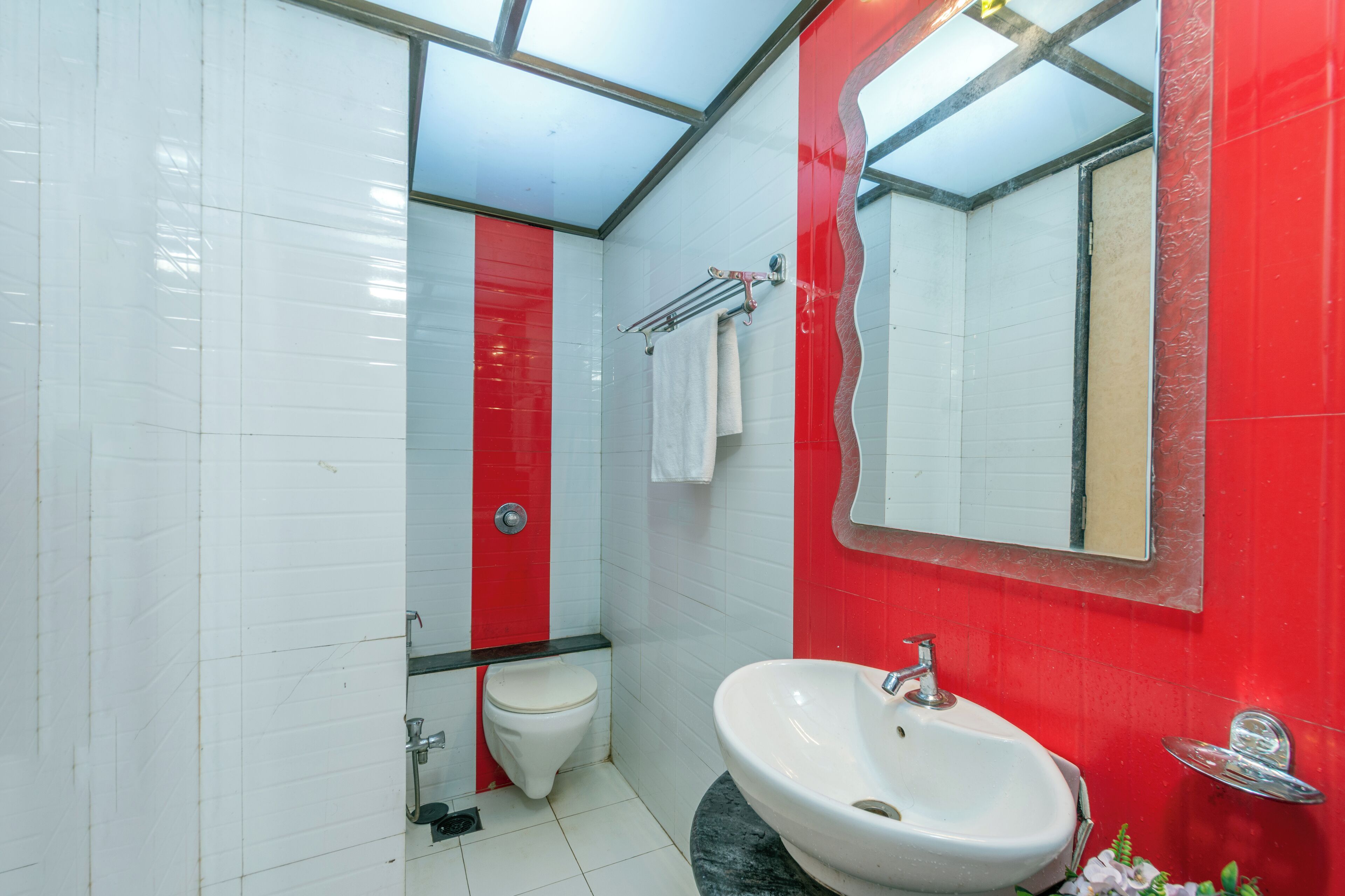 Et badeværelse med vægge i hvid og rød flise, et hvidt toilet, en hvid vask med kromarmatur og et spejl med rød ramme.