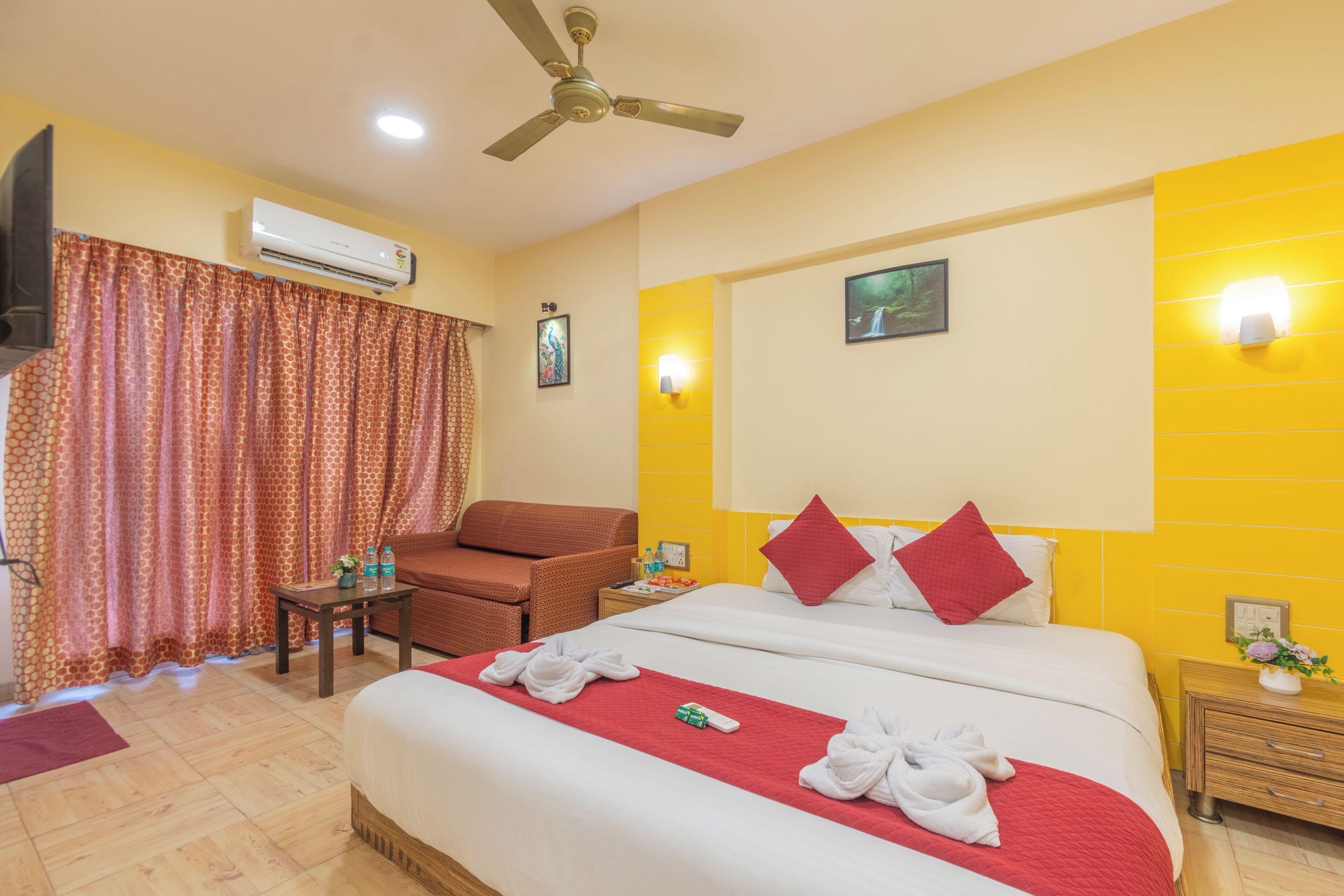 A hotel room with a bed, a chair, a small table, an air conditioner, a ceiling fan, and a window with curtains.