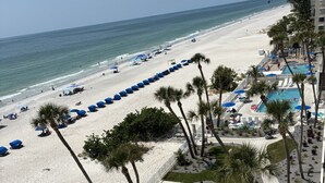 A beach with rows of blue umbrellas, palm trees, and a pool area with lounge chairs.