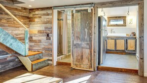 A rustic interior with a wooden staircase, a sliding door leading to a kitchen, and a view of the outside through a window. - Historic Nature Lovers Cabin on Farm in Paonia! (Paonia)
