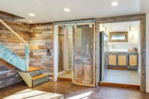 Un interior rústico con una escalera de madera, un acceso corredizo hacia una cocina y vistas al exterior a través de una ventana.