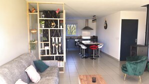 Een moderne woonkamer met een grijze bank, een houten salontafel en een groene fauteuil. In de achtergrond is een keuken zichtbaar met een fornuis, gootsteen en barstoelen.
