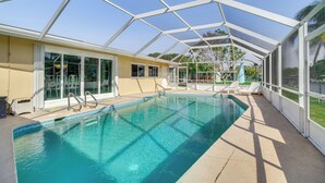 A covered swimming pool with clear blue water, surrounded by a white frame structure, and a view of a green lawn and palm trees in the background. - Centrally Located Cocoa Beach Haven: Walk to Shore (Cocoa Beach)