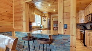 A wooden interior with a stone backsplash, wooden bar stools, and a kitchen area with a microwave and stove. - The Toasted Marshmallow Ski & Lake Game Barn (Canandaigua)