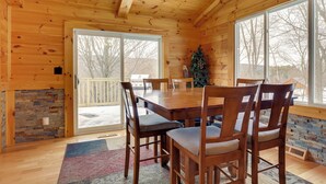 A wooden cabin interior with a dining table, chairs, a fireplace, and large windows. - The Toasted Marshmallow Ski & Lake Game Barn (Canandaigua)