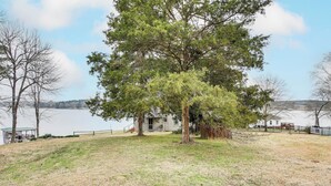 A house with a large tree in front, a grassy yard, and a body of water in the background. - Private Dock + Porch: Serene Lake Greenwood Escape (Greenwood)