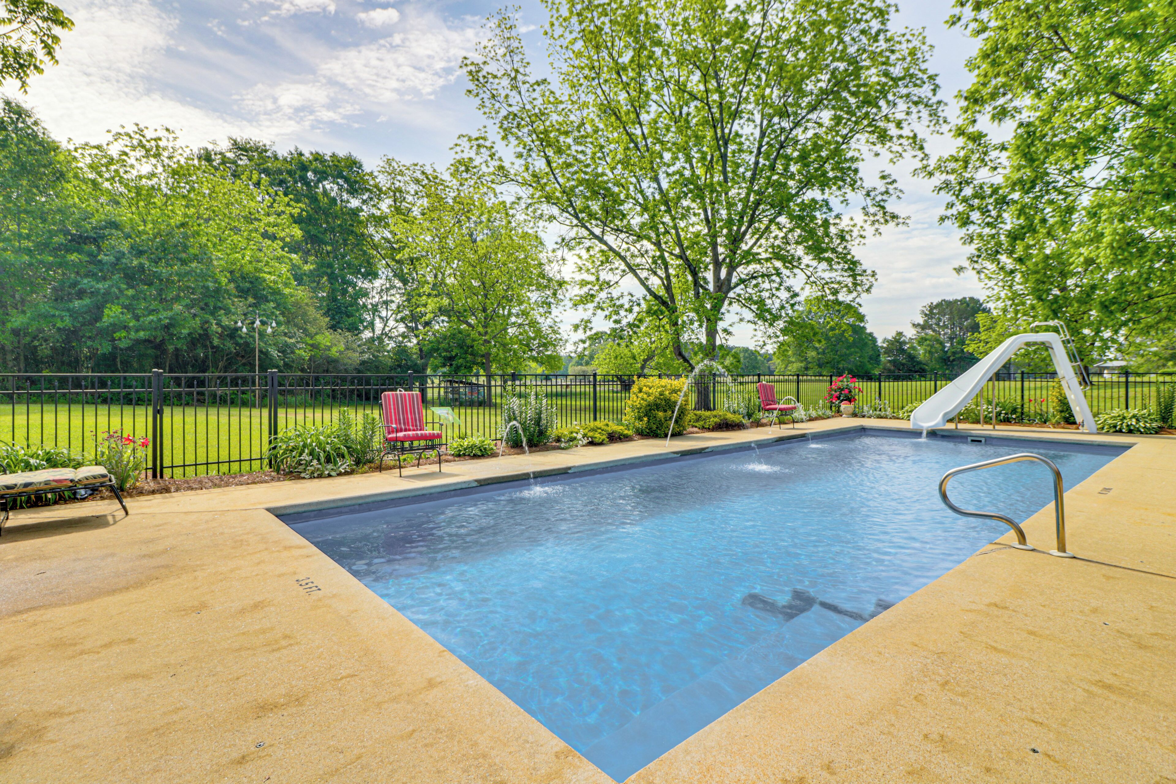 Una piscina rectangular con una escalera metálica y un tobogán, rodeada por un deck de concreto y una cerca de metal negro.