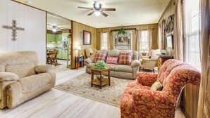 A living room with a beige sofa, a floral armchair, and a wooden coffee table. A ceiling fan is visible, and there is a kitchen in the background. - Yellow Home in Lineville on 2 Acres w/ Pool Access (Lineville)