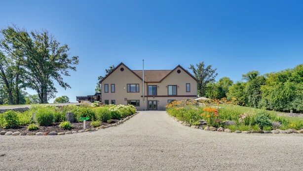 A two-story building with a symmetrical facade, a gravel driveway leading to the entrance, and landscaped gardens with various plants and flowers on either side. - 10-acre Plot: Peaceful Getaway in Webster! (Webster)