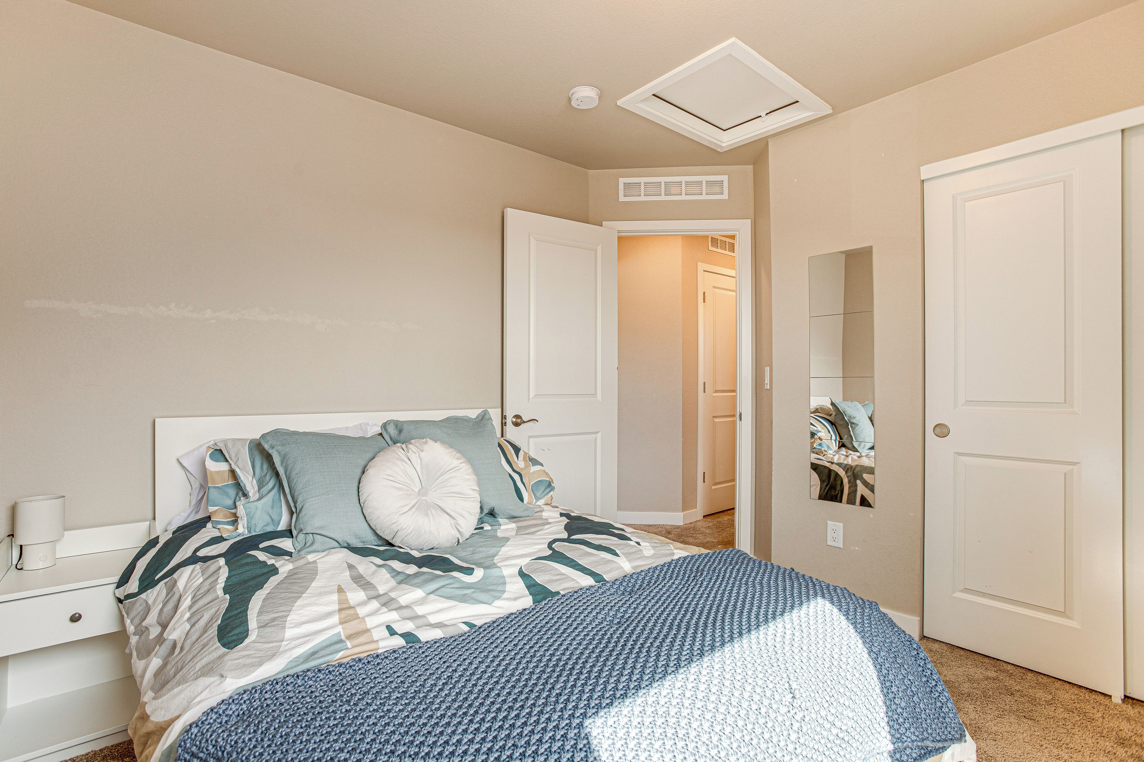 A bedroom with a bed, a nightstand, a bedside table, a door, and a ceiling vent.