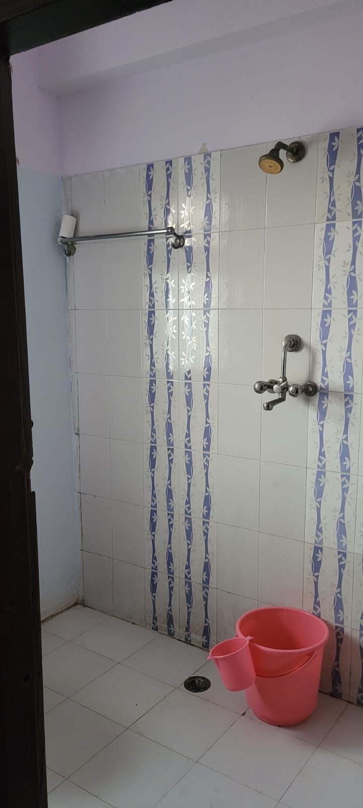 Zona de ducha con pared de azulejos, ducha, alcachofa de ducha manual y un cubo rosa en el suelo.