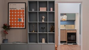 Ein modernes Interieur mit einer grauen Regaleinheit, die verschiedene dekorative Gegenstände und einen gerahmten Bauhaus-Plakat zeigt, sowie ein Blick in einen Küchenbereich.