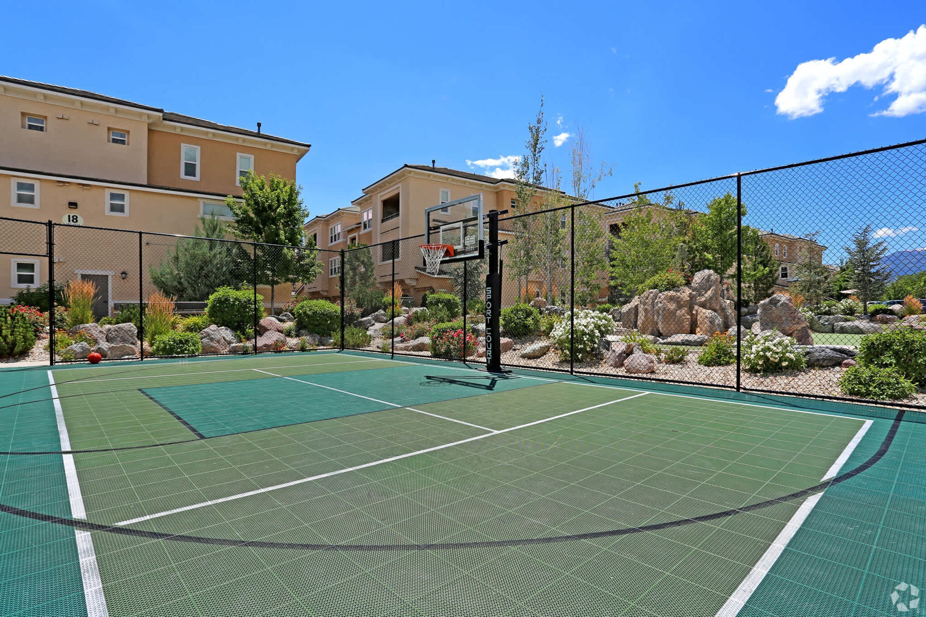 Una cancha de tenis al aire libre con un aro de baloncesto, rodeada por una valla y follaje.