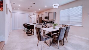 A modern kitchen with white cabinets, a central island, and a dining area with a marble table and velvet chairs. - 15976 Spacious Charming Family Getaway (Clermont)