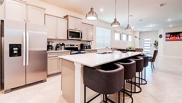 A modern kitchen with stainless steel appliances, a central island with bar stools, pendant lighting, and a dining area with a view. - 15976 Spacious Charming Family Getaway (Clermont)