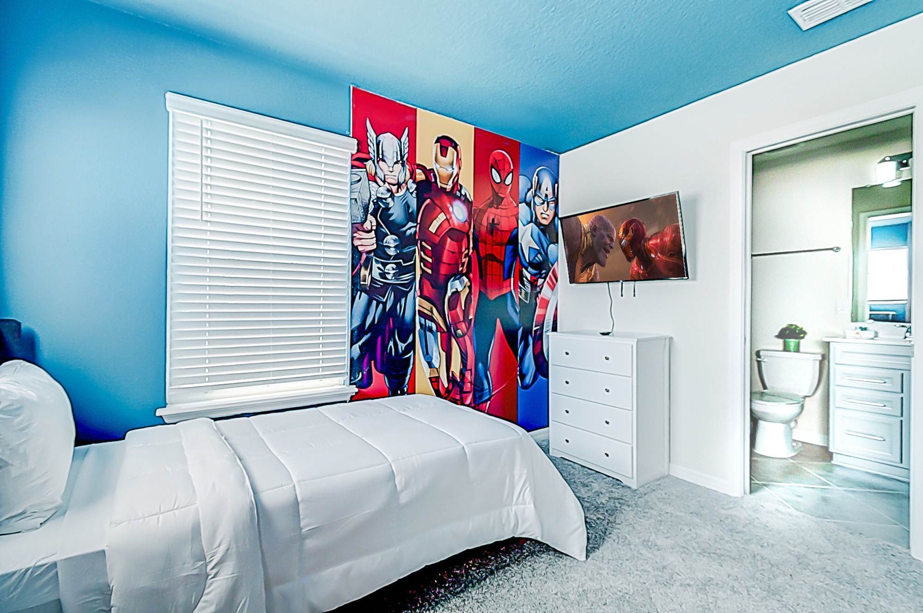 Quarto com uma cama grande, penteadeira branca, televisão na parede e banheiro visível por uma porta aberta.