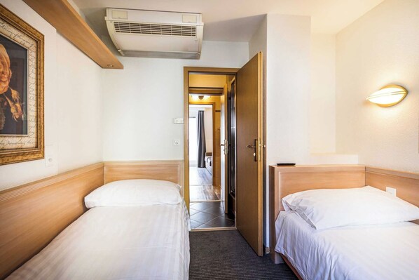 A hotel room with two single beds, a wooden headboard, a wall-mounted air conditioner, a framed picture, and a bedside lamp.
