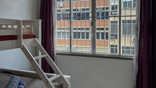 A room with a bunk bed, a ceiling fan, a window with curtains, and a view of a building outside.