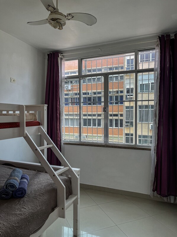 A room with a bunk bed, a ceiling fan, a window with curtains, and a view of a building outside.