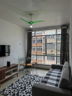 A living room with a sofa, a TV, a ceiling fan, a dining table with chairs, and large windows with curtains.
