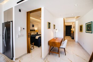 A hotel room with a bed, a desk with a chair, a refrigerator, and a hallway with artwork.