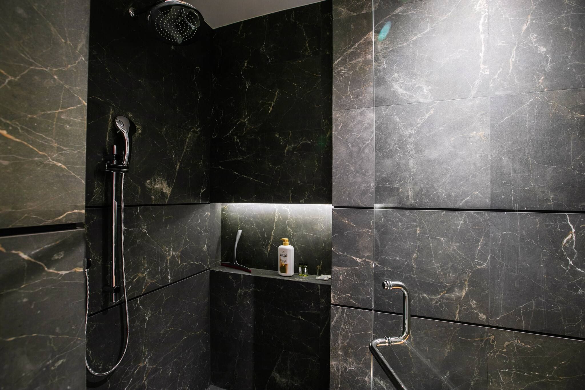 A modern bathroom with a black marble shower, a handheld showerhead, and a glass shelf with toiletries.