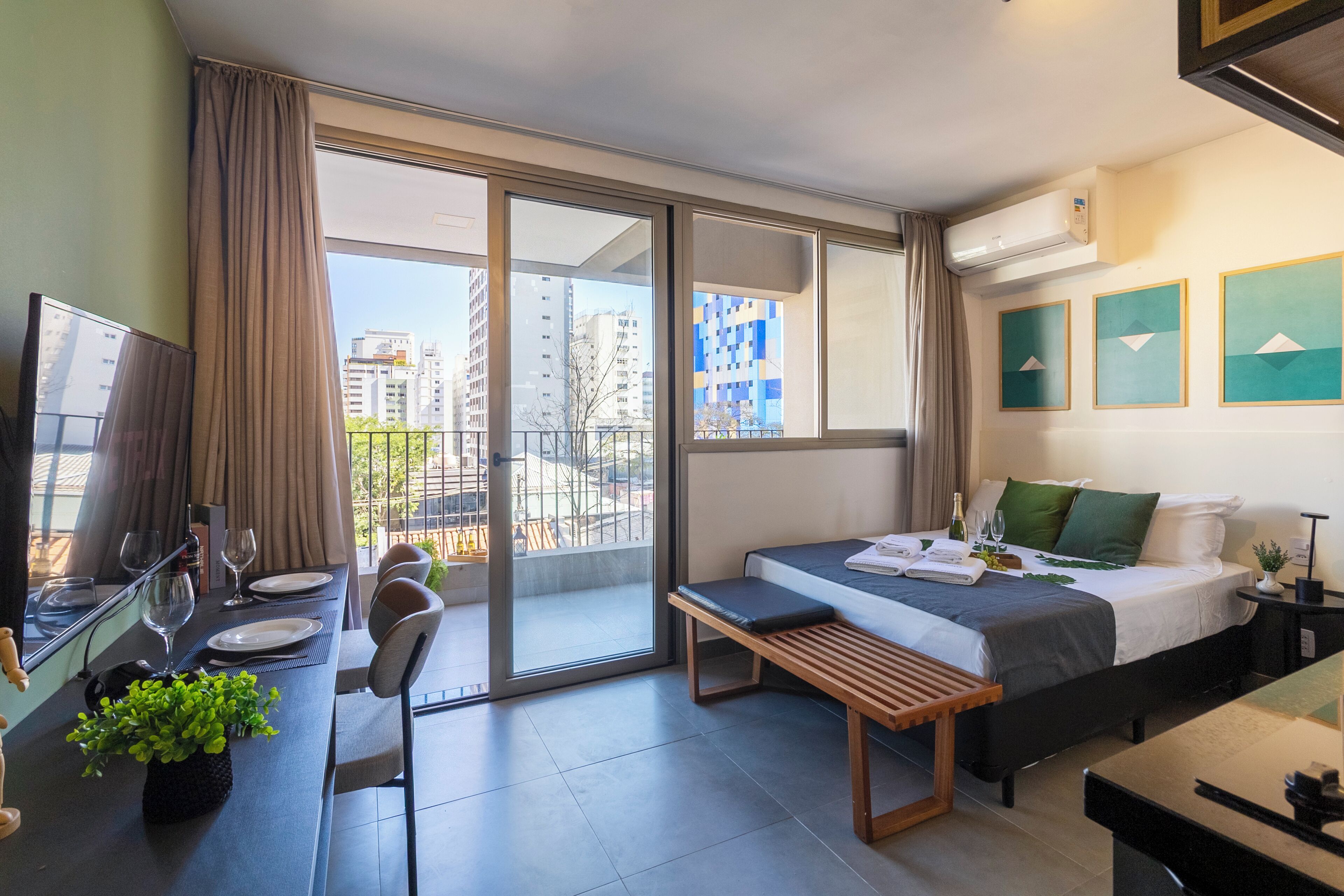 Una habitación de hotel moderna con cama, escritorio con computadora, banca, zona de comedor y balcón con vista a la ciudad.