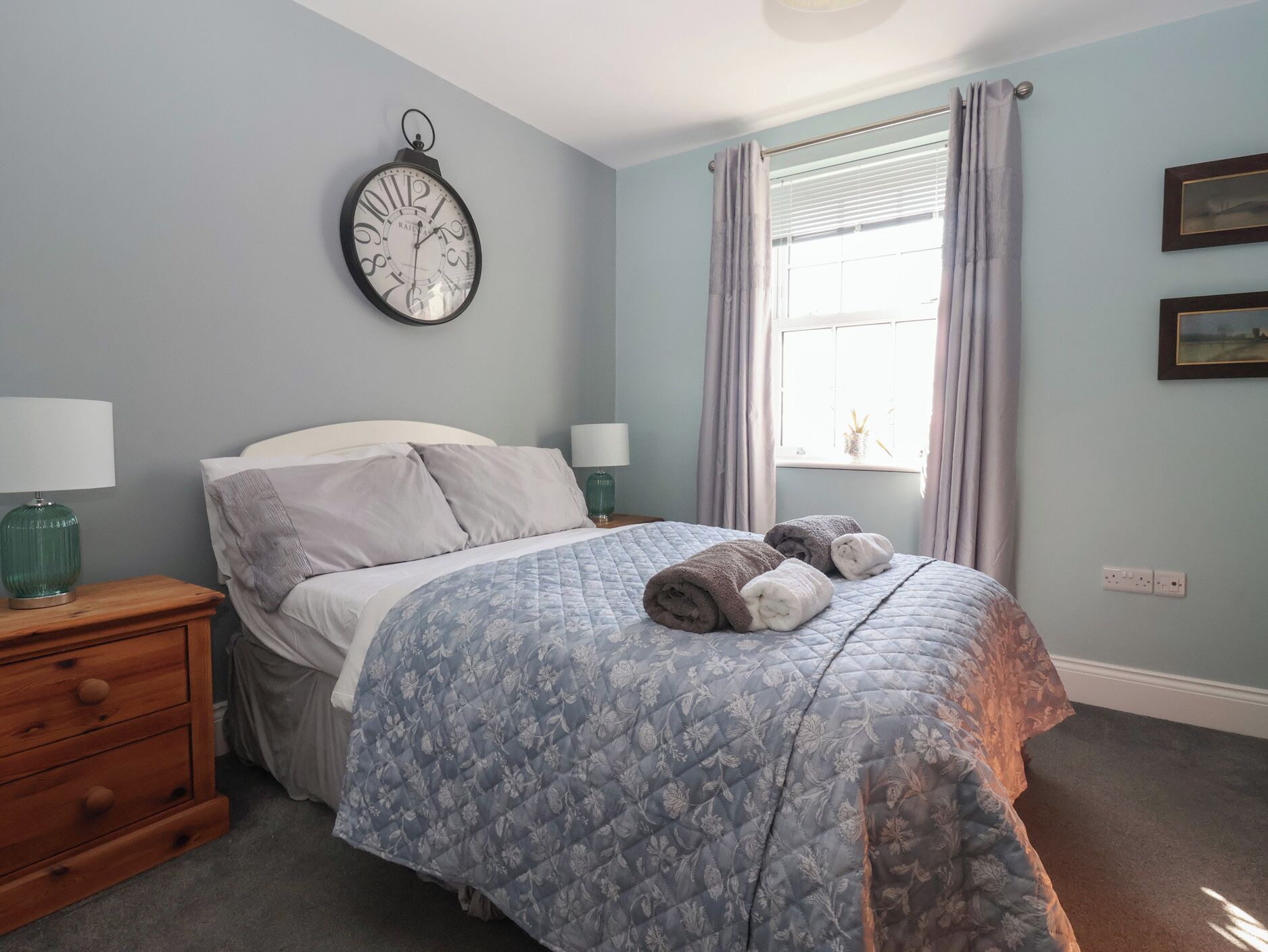 A bedroom with a bed, a nightstand, a wall clock, a window with curtains, and a wall outlet.
