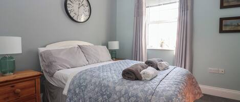 A bedroom with a bed, a nightstand, a wall clock, a window with curtains, and a wall outlet.