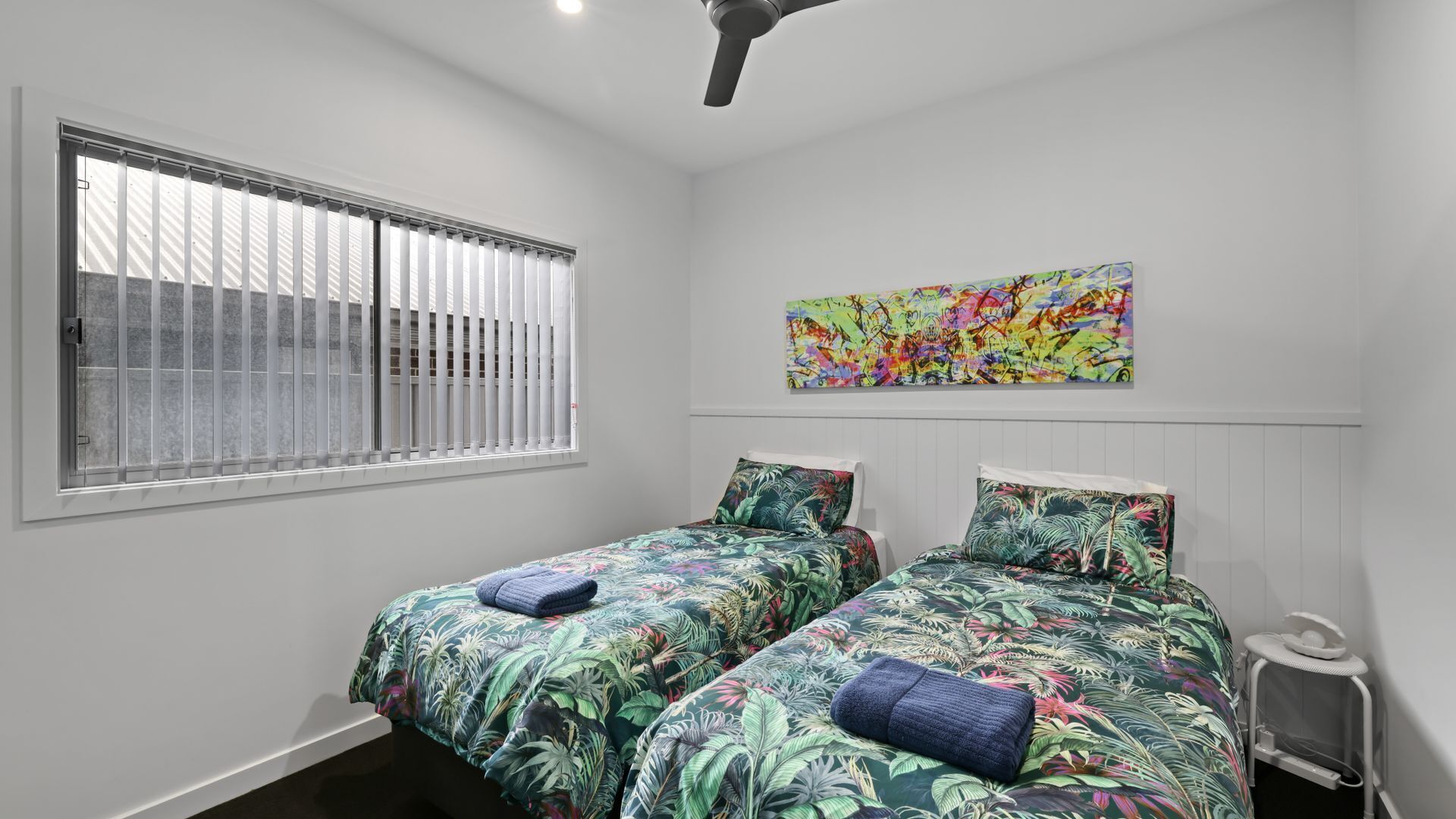 A bedroom with two beds, a window with blinds, a ceiling fan, and a colorful abstract painting on the wall.