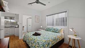 A bedroom with a bed, a bedside table with a lamp, a window with blinds, a door, and a view of the kitchen. - Secret on Seagrass by Book Jervis Bay (Vincentia)