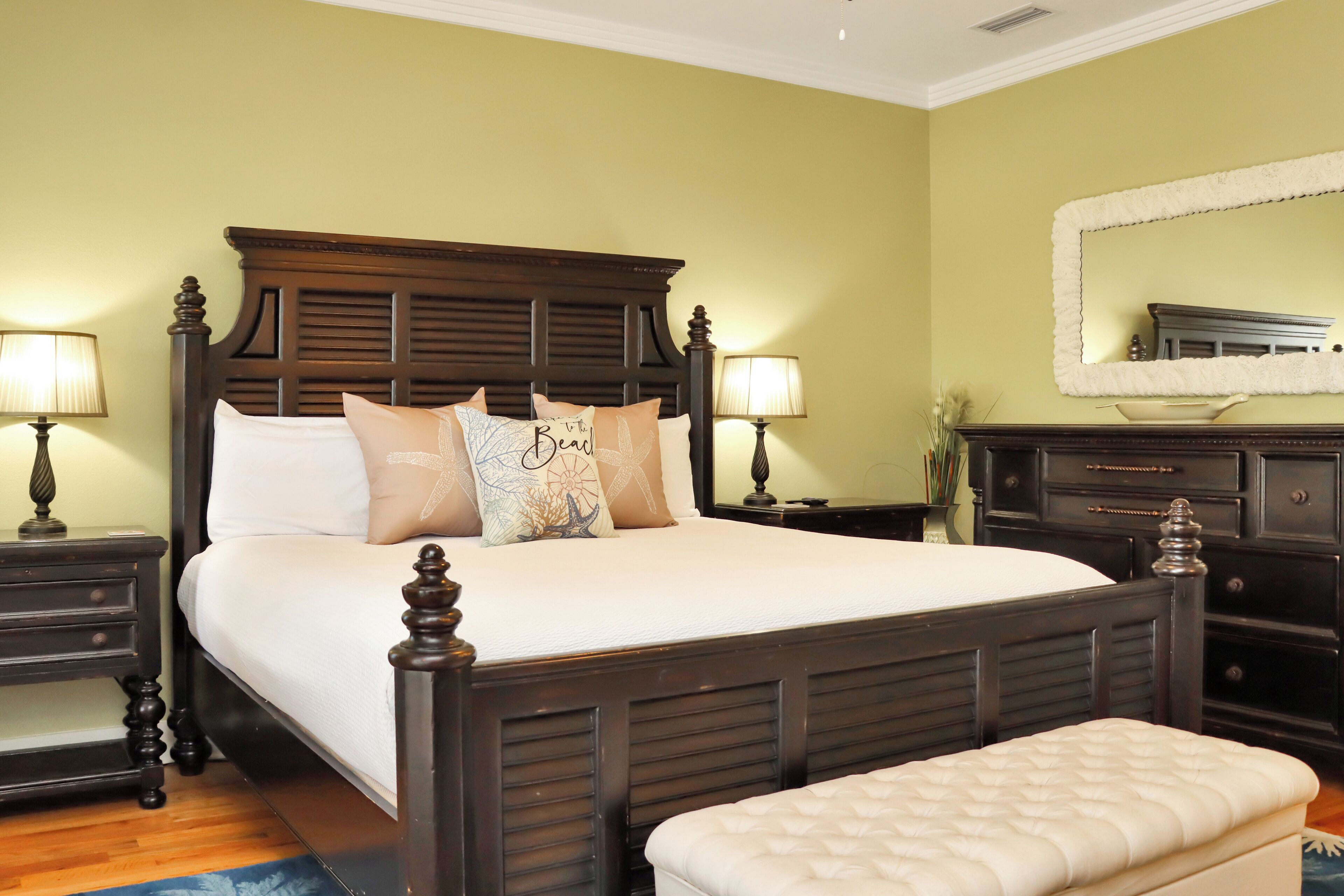 A neatly made bed with a wooden headboard, two bedside tables with lamps, a large mirror above a dresser, and a bench at the foot of the bed.