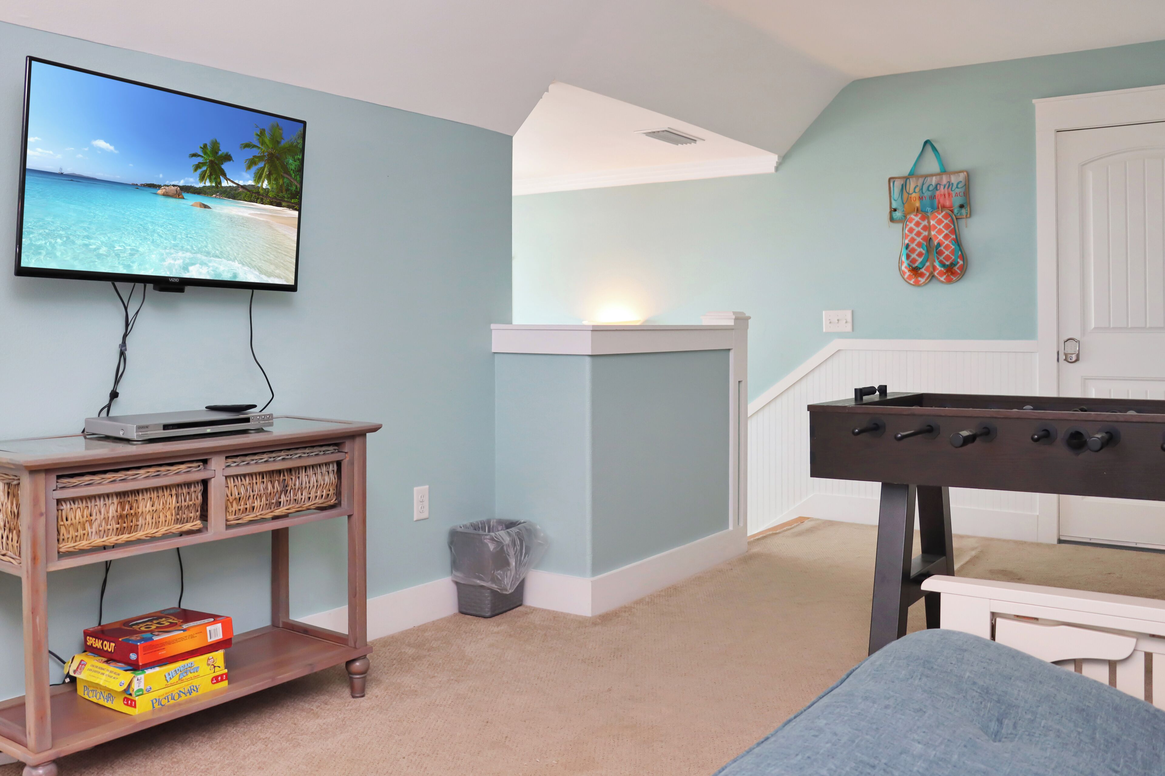 A room with a television on a wooden stand, a foosball table, and a wall-mounted welcome sign.