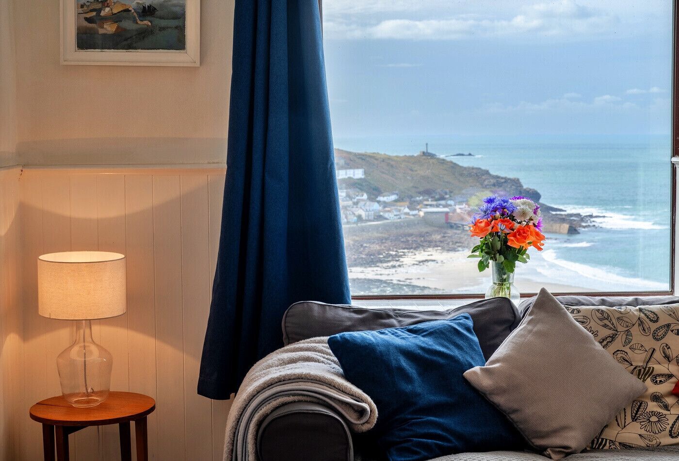 A room with a view of the ocean, a lamp, a vase with flowers, and a patterned pillow.