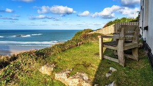 A wooden bench by a coastal path with a view of the ocean and a building in the background. - Bishop Rock (Sennen)