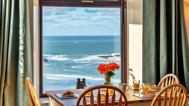 A dining table with a view of the ocean through a large window, a vase with red flowers, and breakfast items including croissants and orange juice. - Bishop Rock (Sennen)