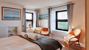 A hotel room with a bed, two wicker chairs, a small table, a shelf with decorative items, and a painting on the wall. - Bishop Rock (Sennen)
