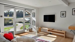 A modern living room with a large sliding glass door leading to a garden, a flat-screen TV mounted on the wall, and a wooden entertainment unit.